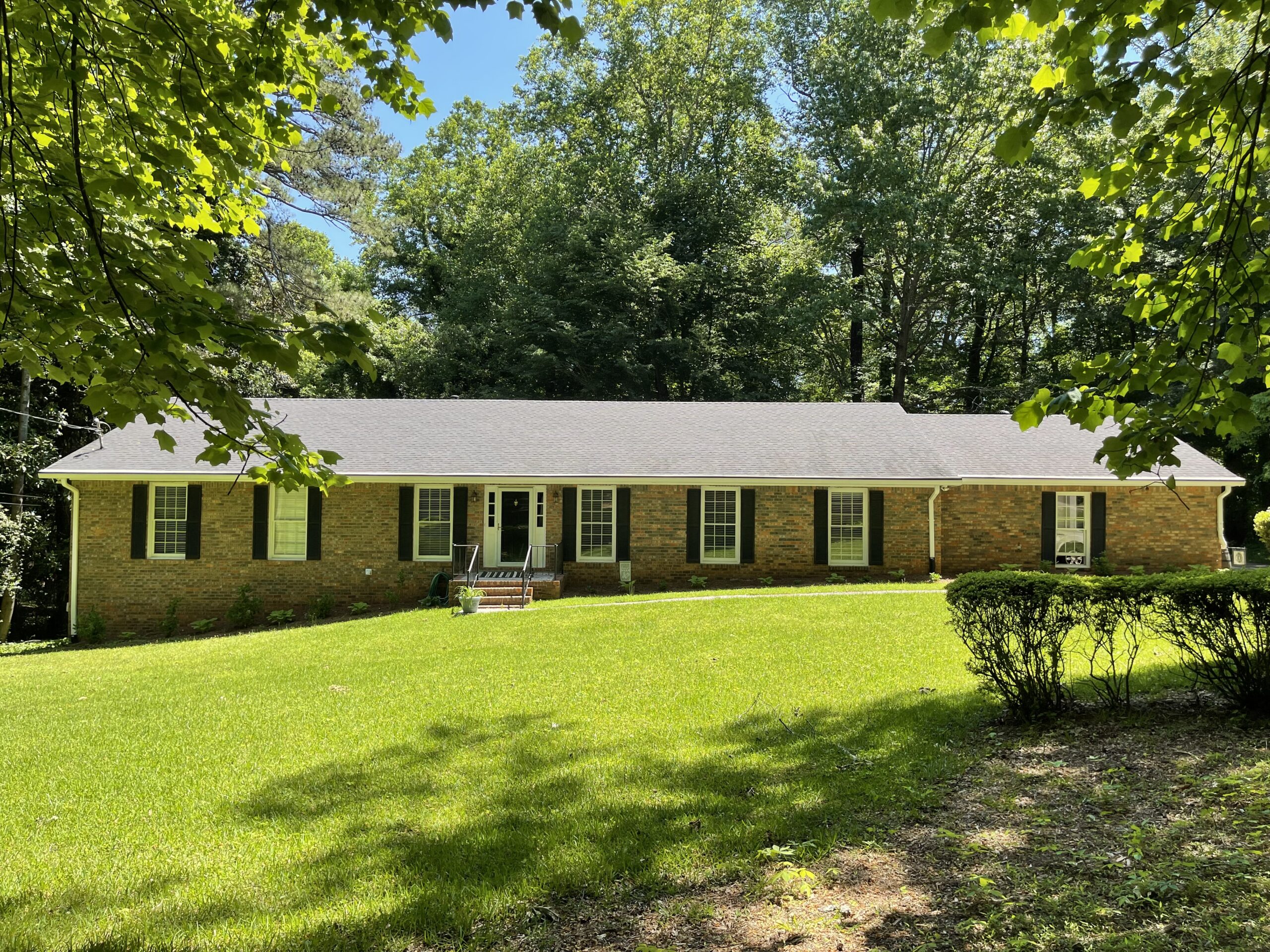 Single-story brick house with black shutters, front yard, and large trees framing the view on a sunny day.