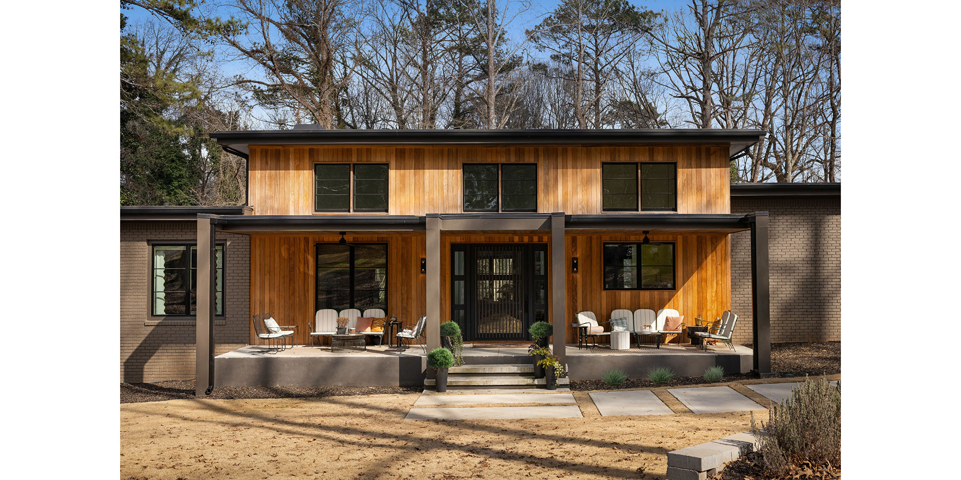 Modern two-story house with vertical wood siding and brick accents, a covered front porch with outdoor seating, and a landscaped yard with trees in the background.