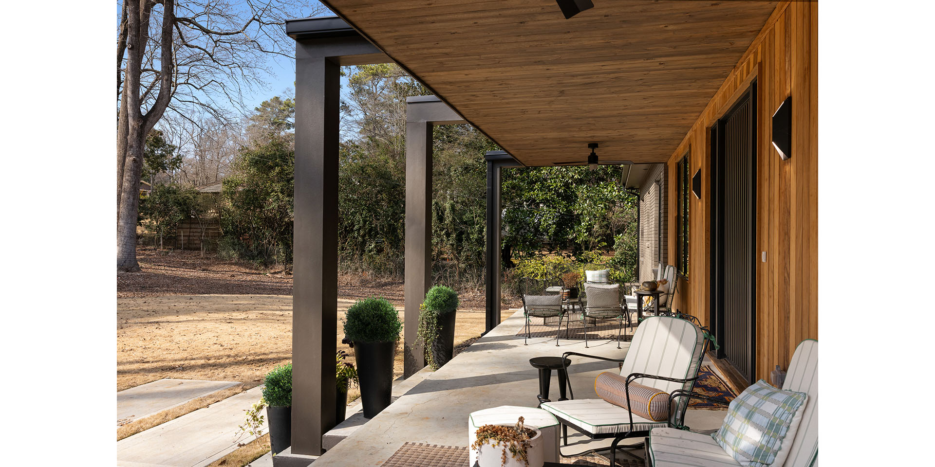Covered patio with wooden ceiling and metal posts; outdoor seating area with wicker chairs and cushions along a brick house, yard beyond