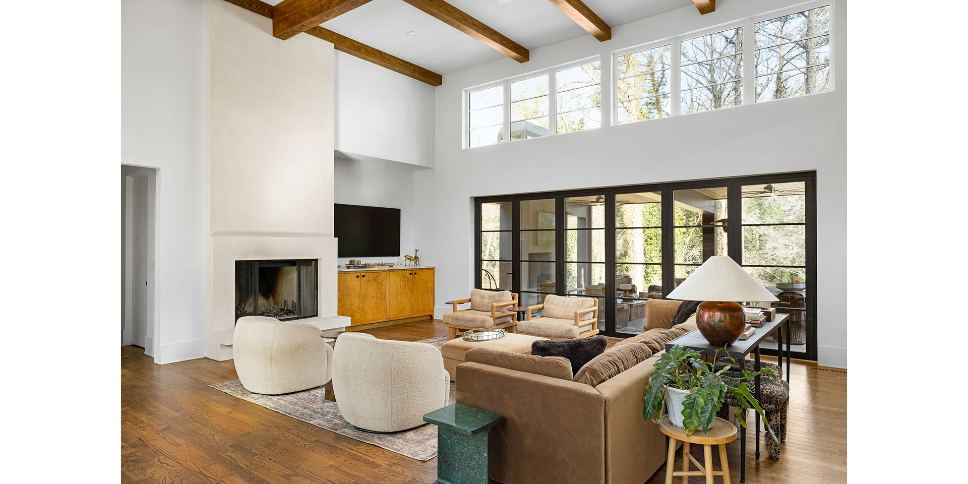Bright, modern living room with high ceilings, exposed wooden beams, and large windows overlooking greenery.