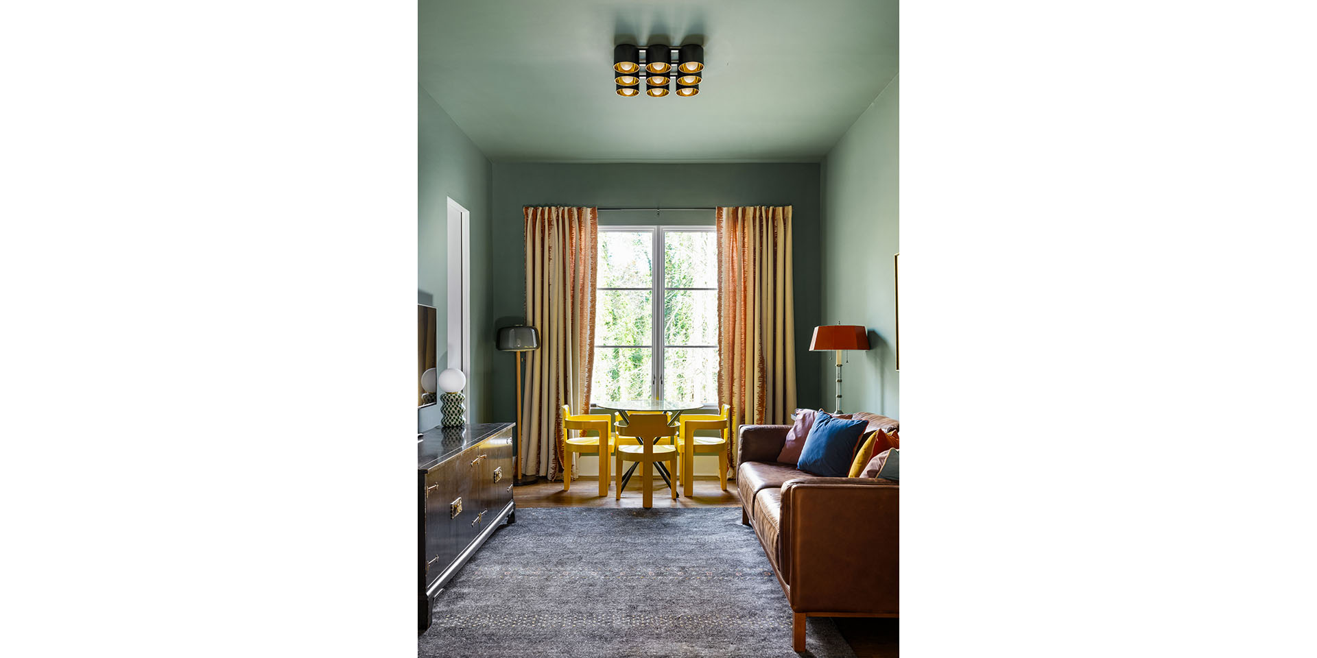 Living room with green walls, a window with striped curtains, and a small yellow dining table and chairs in front of it.