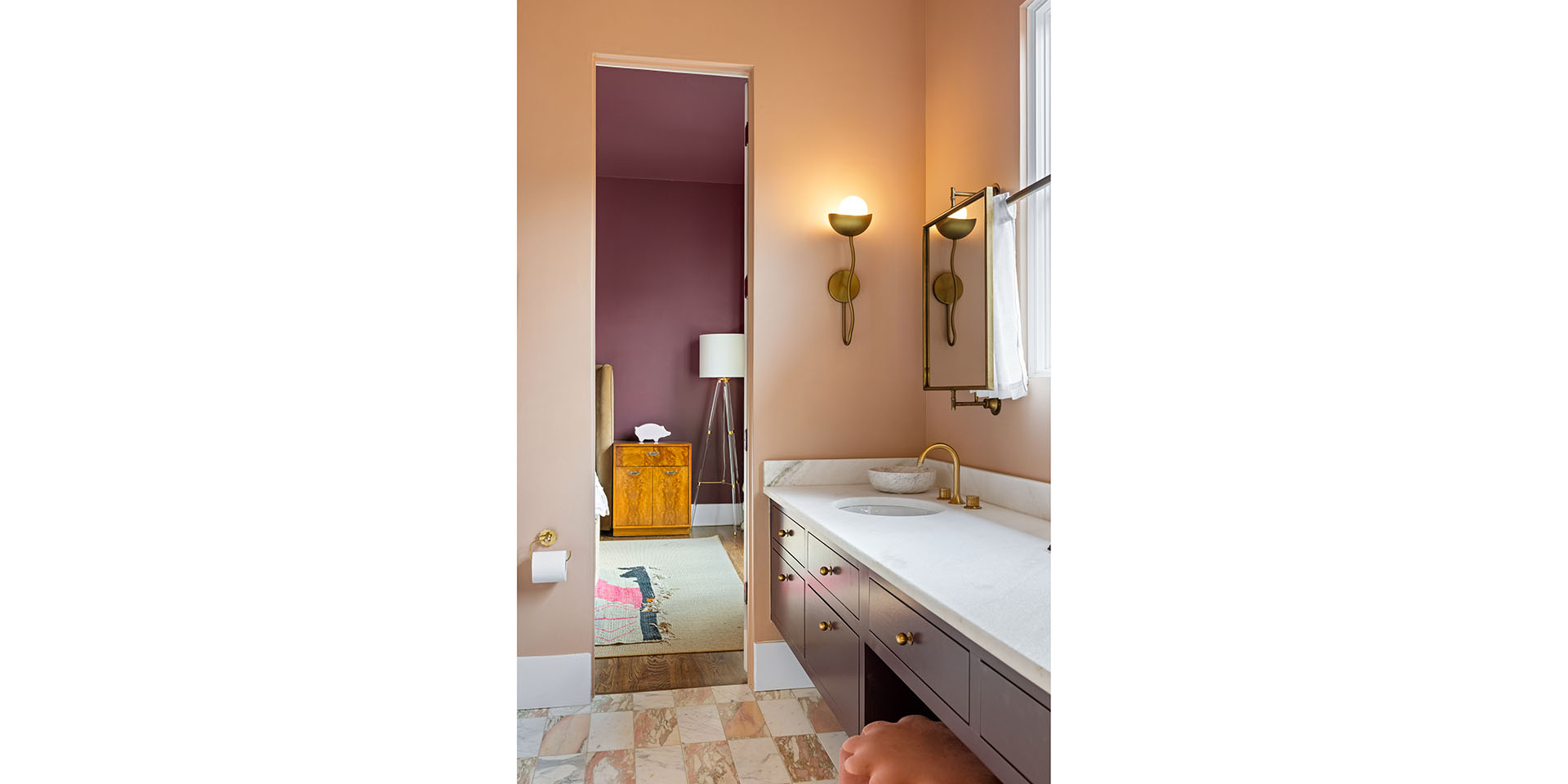 Bathroom with peach walls and marble counter, brass faucet and towel bar; doorway opens to a purple bedroom with a wooden chest and floor lamp; tiled floor.
