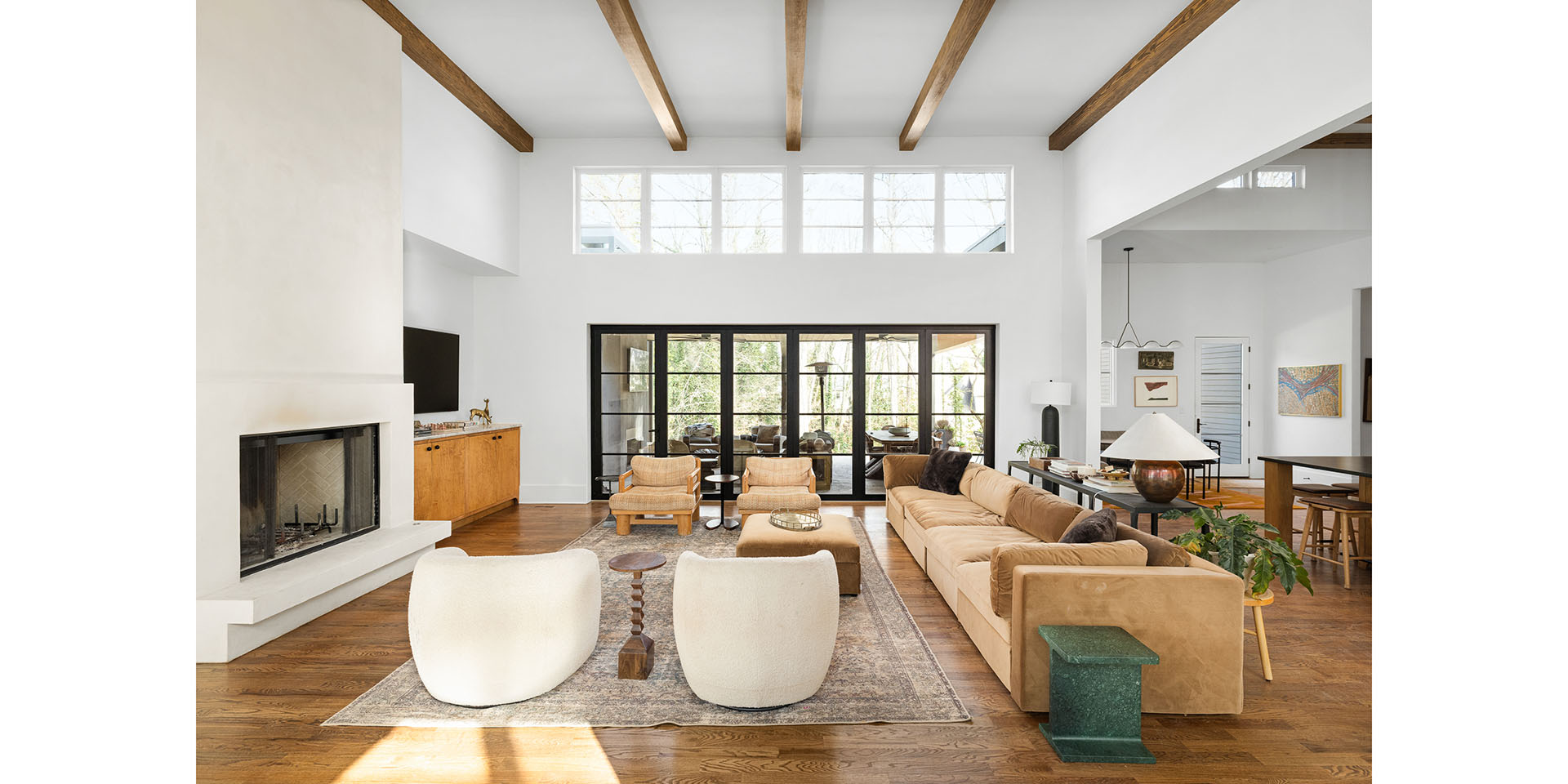 Spacious living room with white walls, exposed wooden beams, and large black-framed glass doors opening to an outdoor area; tan sofa and two cream chairs arranged around a rug.