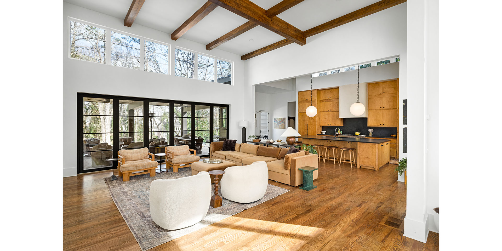 Open-concept living area with hardwood floors, beige sofa and white lounge chairs near a kitchen island with wooden cabinets and stools.|Informative image with a kitchen and seating area in a bright, spacious home.