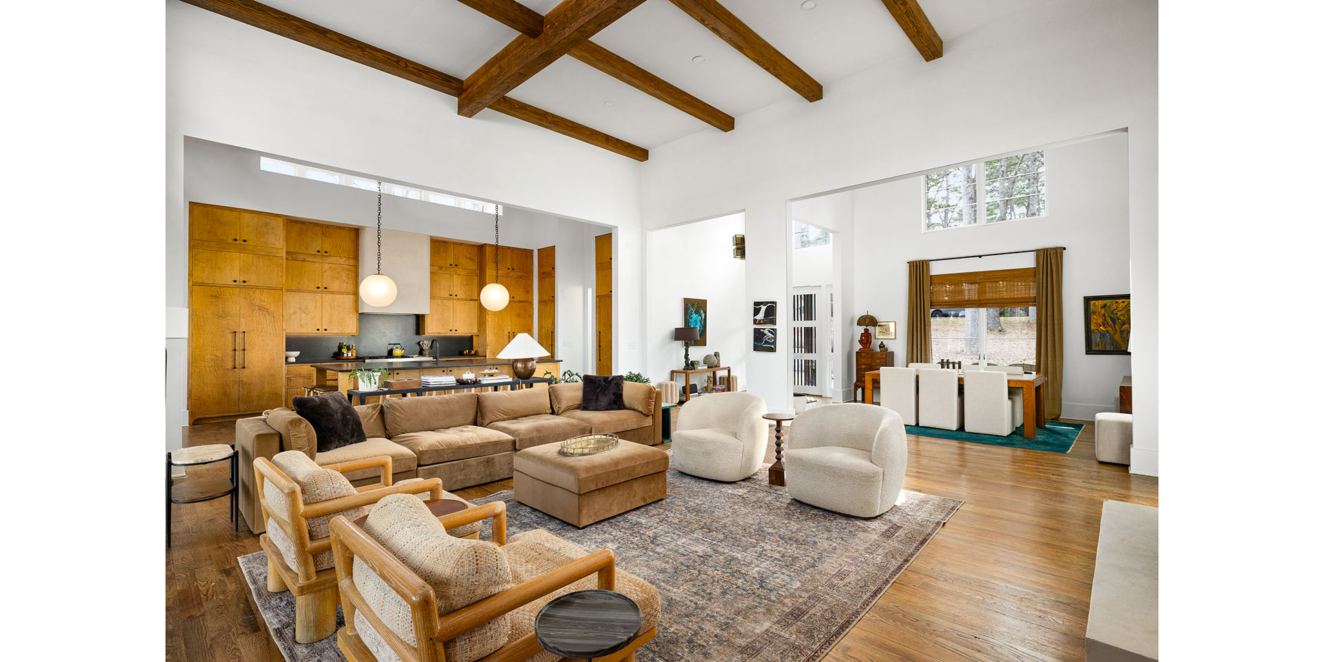 Open-concept living room with exposed wooden ceiling beams, beige sofa, and mid-century chairs near the kitchen.