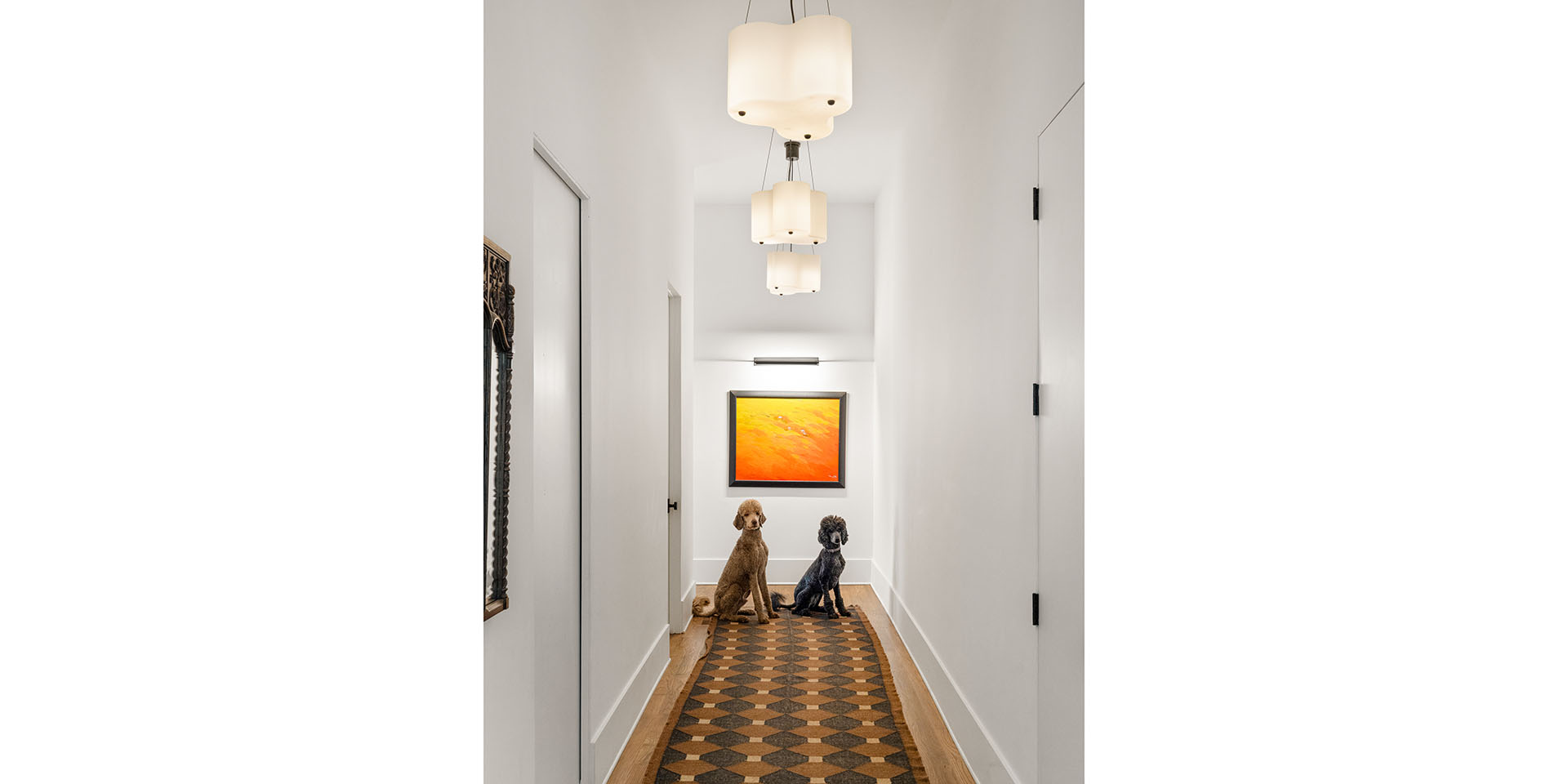 Two dogs sit in a bright, narrow hallway with a geometric tile rug and an orange abstract painting at the far end.