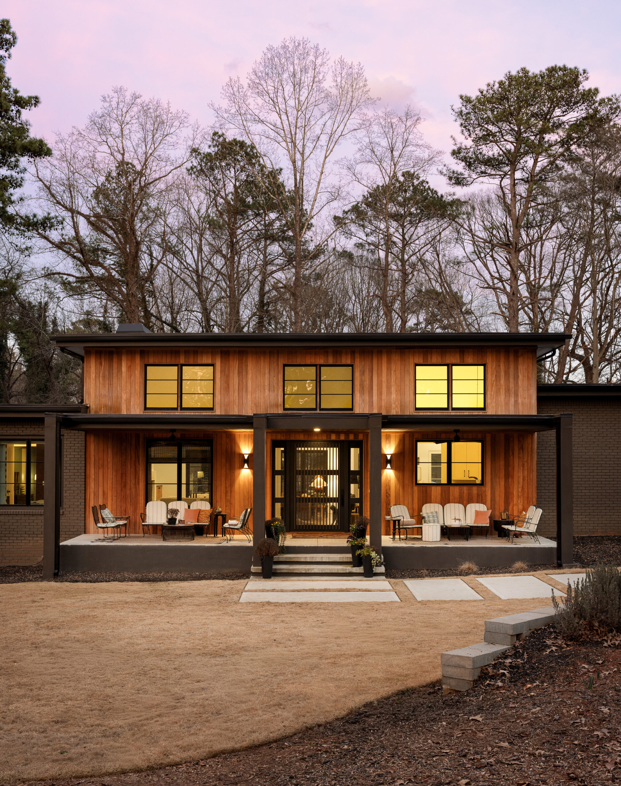 Modern wooden-clad house with a covered porch and yellow-tinted windows, outdoor seating, and leafless trees against a pastel, sunset sky behind.