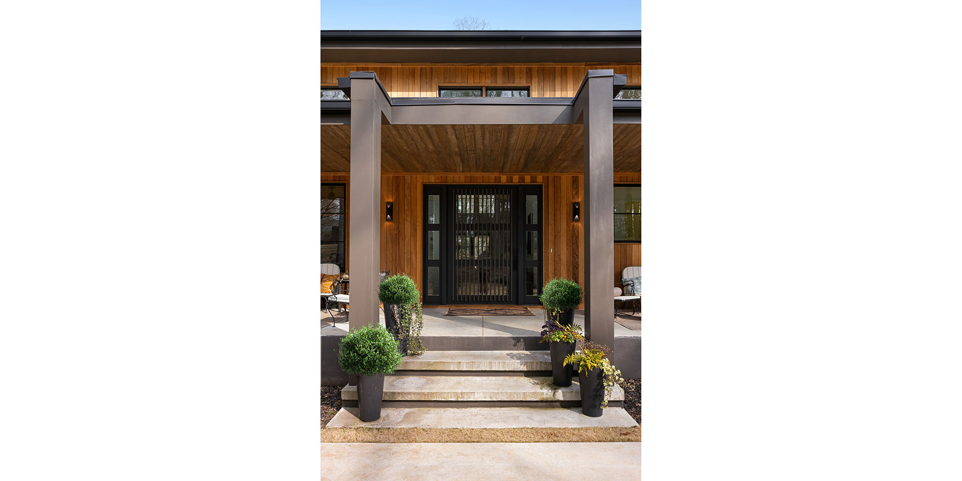 Modern wooden house entrance with a covered porch, black-framed glass door, and potted topiary on concrete steps