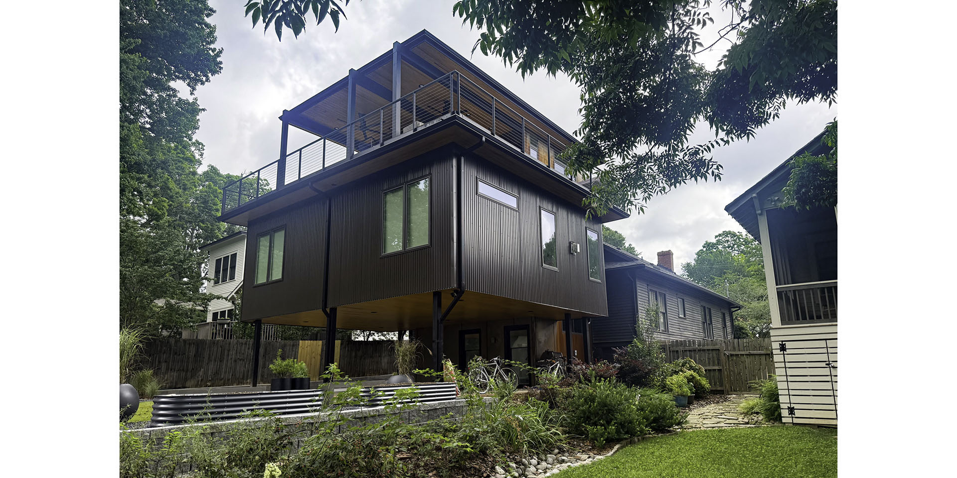 Modern elevated two-story house with a large upper deck, dark metal siding, and surrounding garden in a suburban yard.