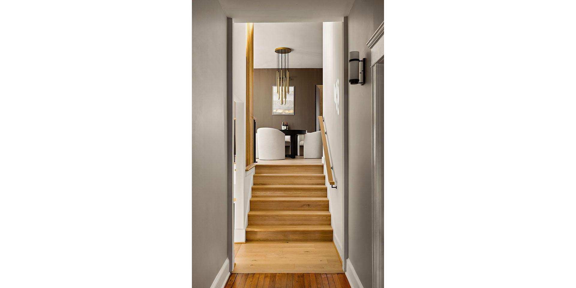 Wood staircase leading up to a dining area with a round black table and white upholstered chairs, beneath a gold pendant chandelier in a modern interior.