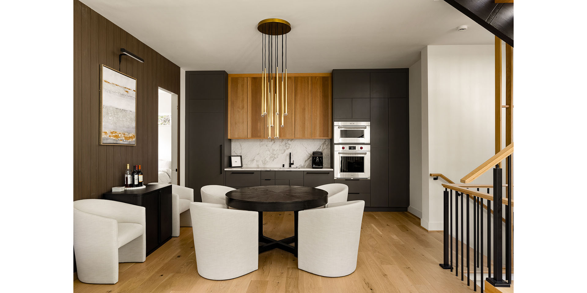 Open-plan kitchen with dark grey cabinets, wooden upper cabinets, marble backsplash, and a round black dining table with white upholstered chairs under a gold pendant light, staircase to the right.
