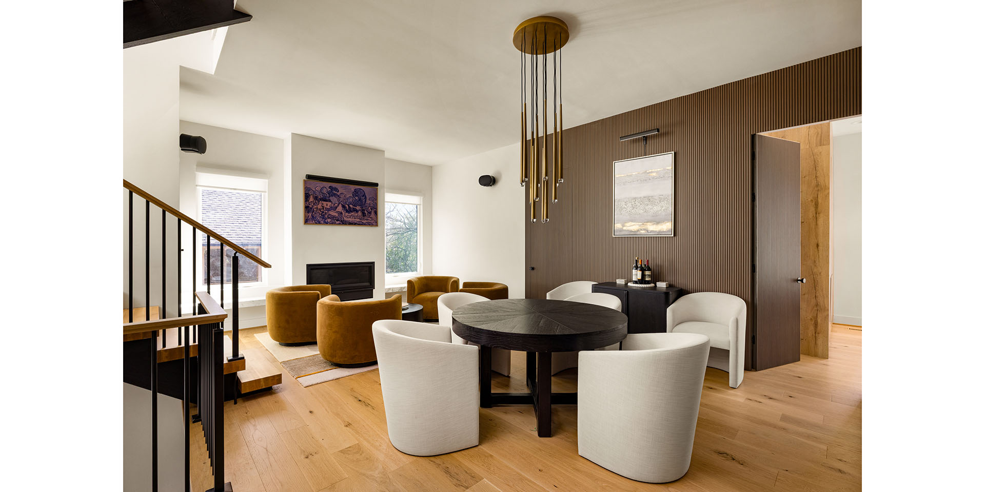 Modern open-concept living/dining area with a round black dining table and light gray upholstered chairs, mustard lounge chairs, and a wood-paneled wall featuring a contemporary gold chandelier.
