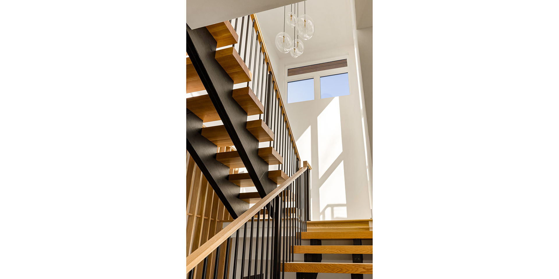 Modern interior staircase with wooden treads and black metal stringers, ascending beside a white wall and large windows with natural light.