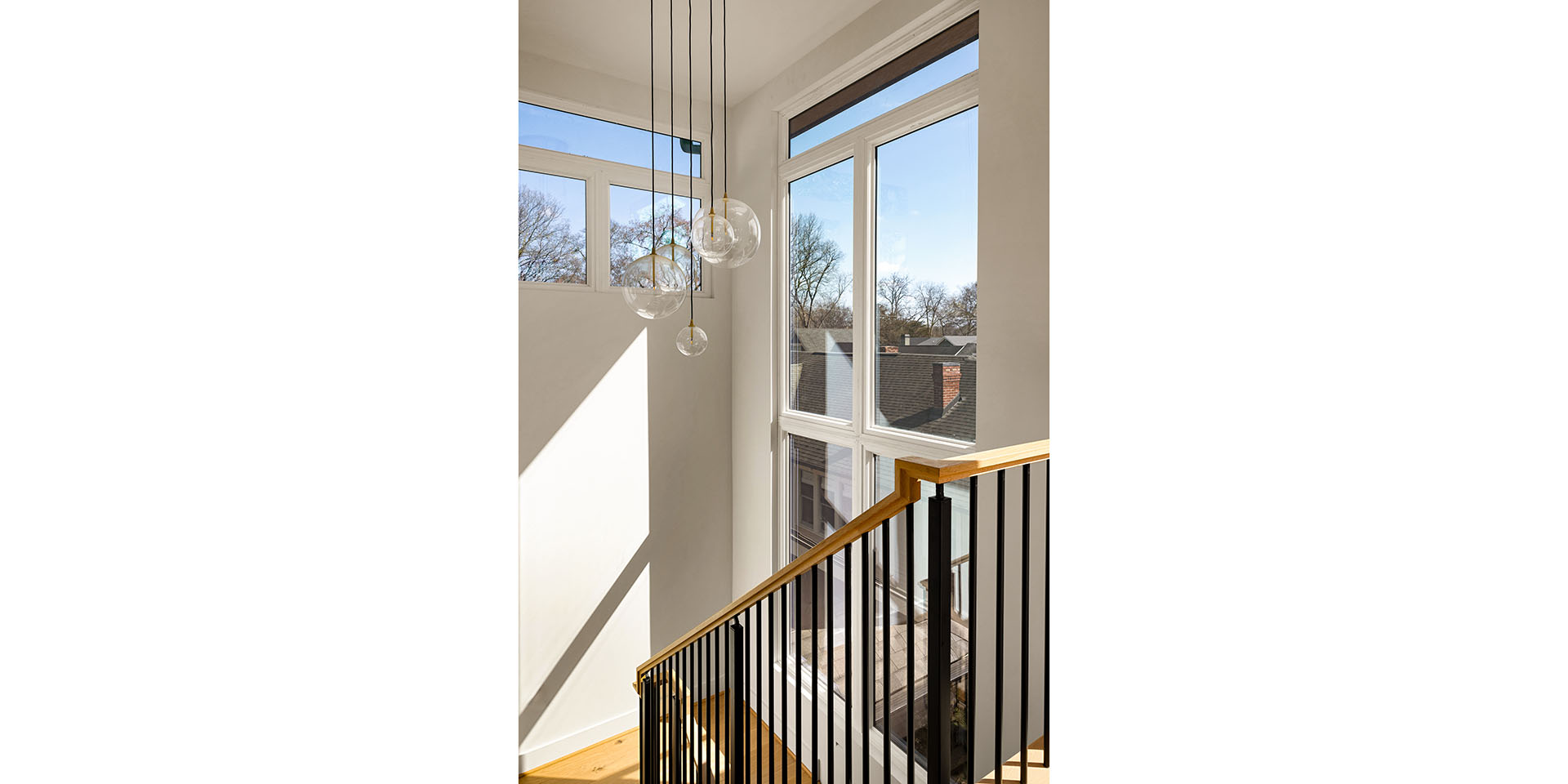 Sunlit stairwell with a wooden handrail, black metal balusters, and hanging glass pendant lights beside large windows.