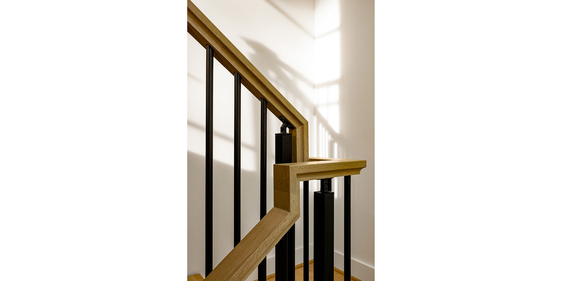 Wooden handrail with black metal balusters along a sunlit staircase, casting shadows on a white wall.