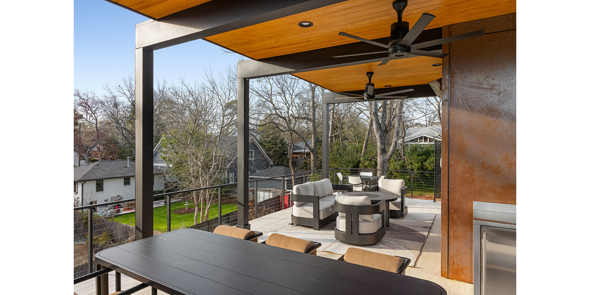 Modern outdoor patio with black metal posts, orange wood ceiling fans, and cream cushioned seating overlooking a suburban yard.