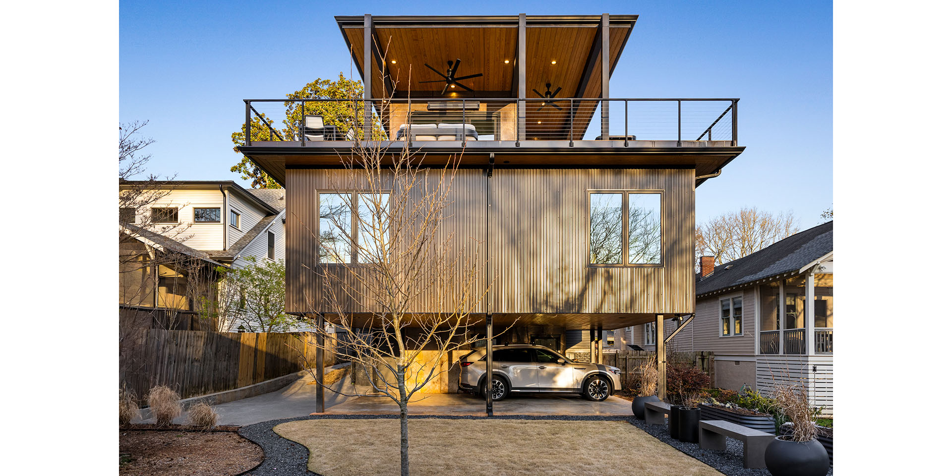 Modern multi-story house with a rooftop deck and glass railing, parked car beneath on a covered level