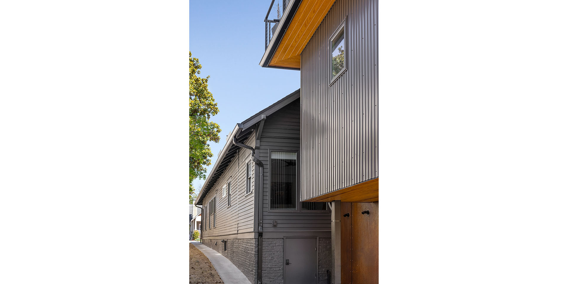 Modern two-story house exterior with gray horizontal siding and metal-clad upper level, small window, and wooden soffit overhangs along a concrete walkway.