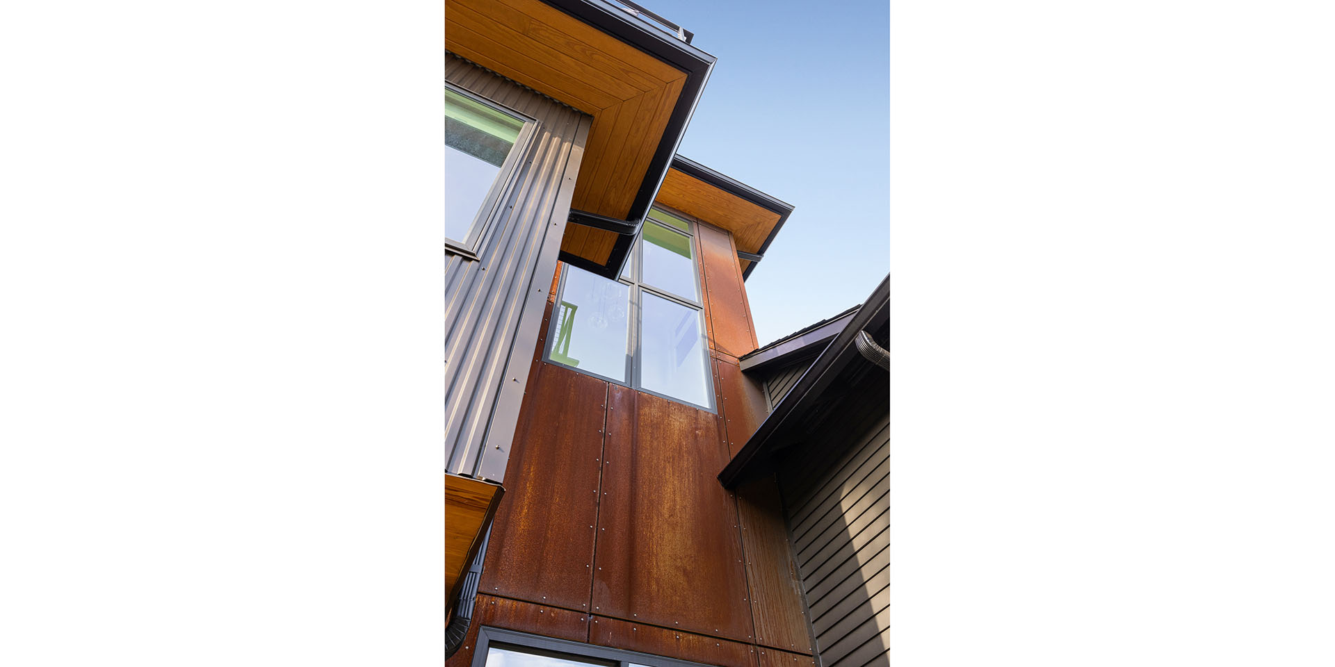 Angular corner of a modern building with rust-colored metal panels, gray siding, and large windows against a blue sky.