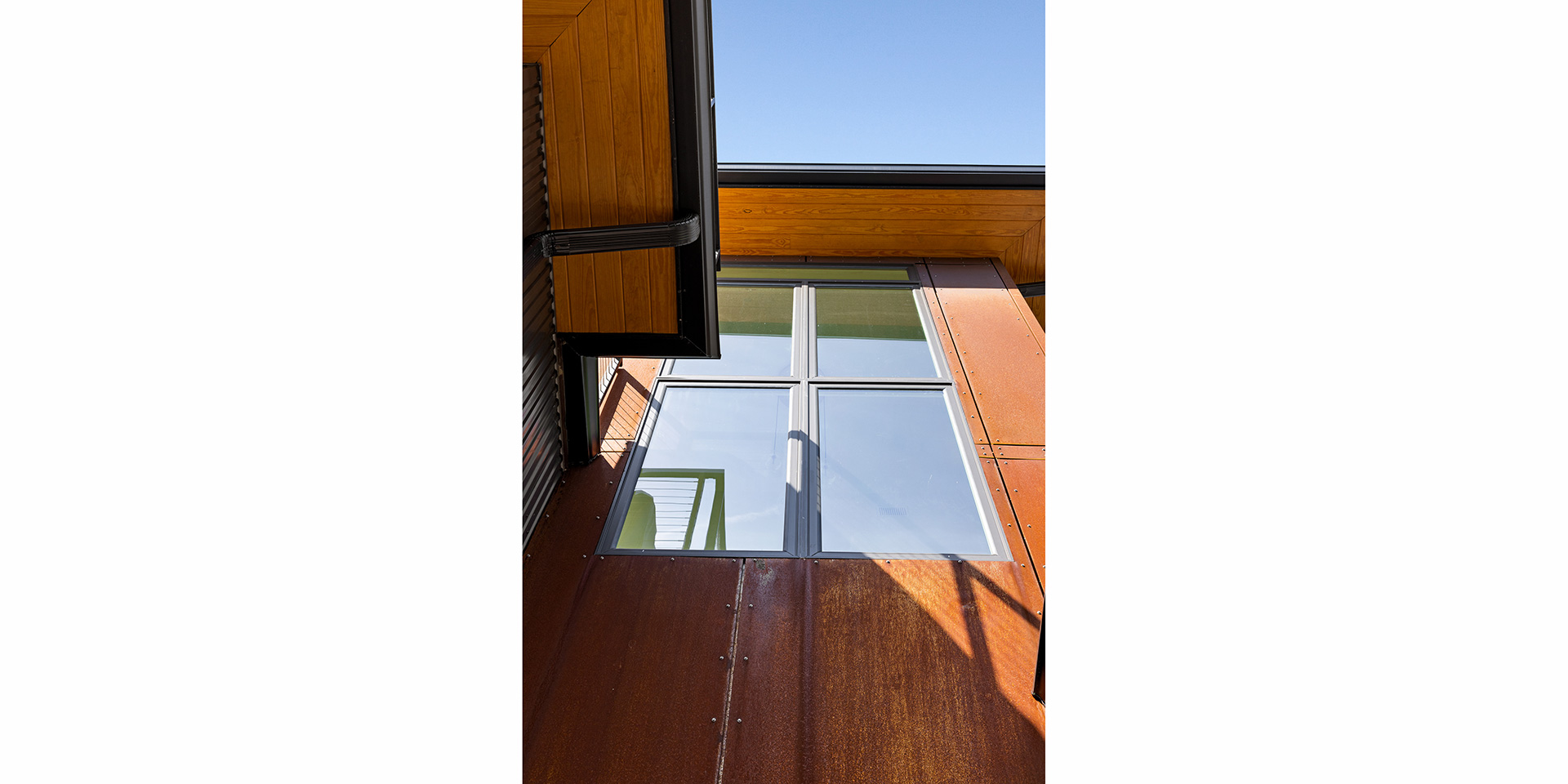 Angular view of a rusty metal wall with a large window and wooden eaves against a blue sky