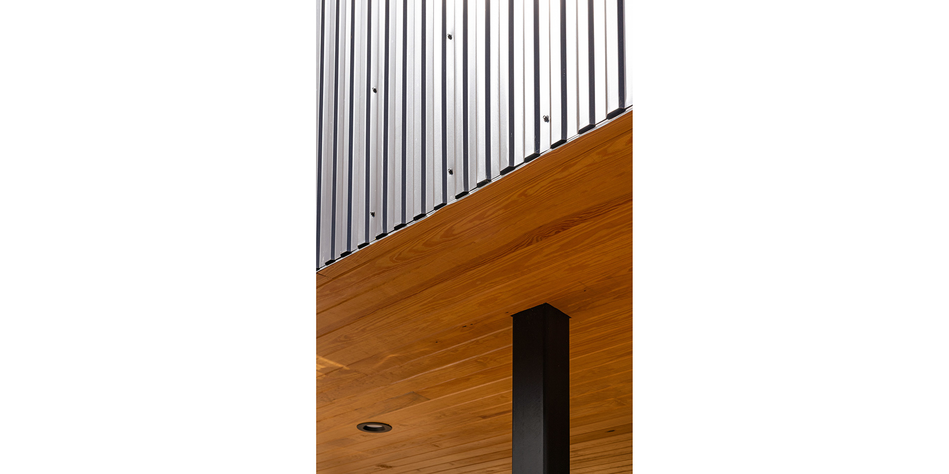 Interior corner showing vertical silver corrugated metal wall panels, warm wooden ceiling, and a black steel post in the foreground.