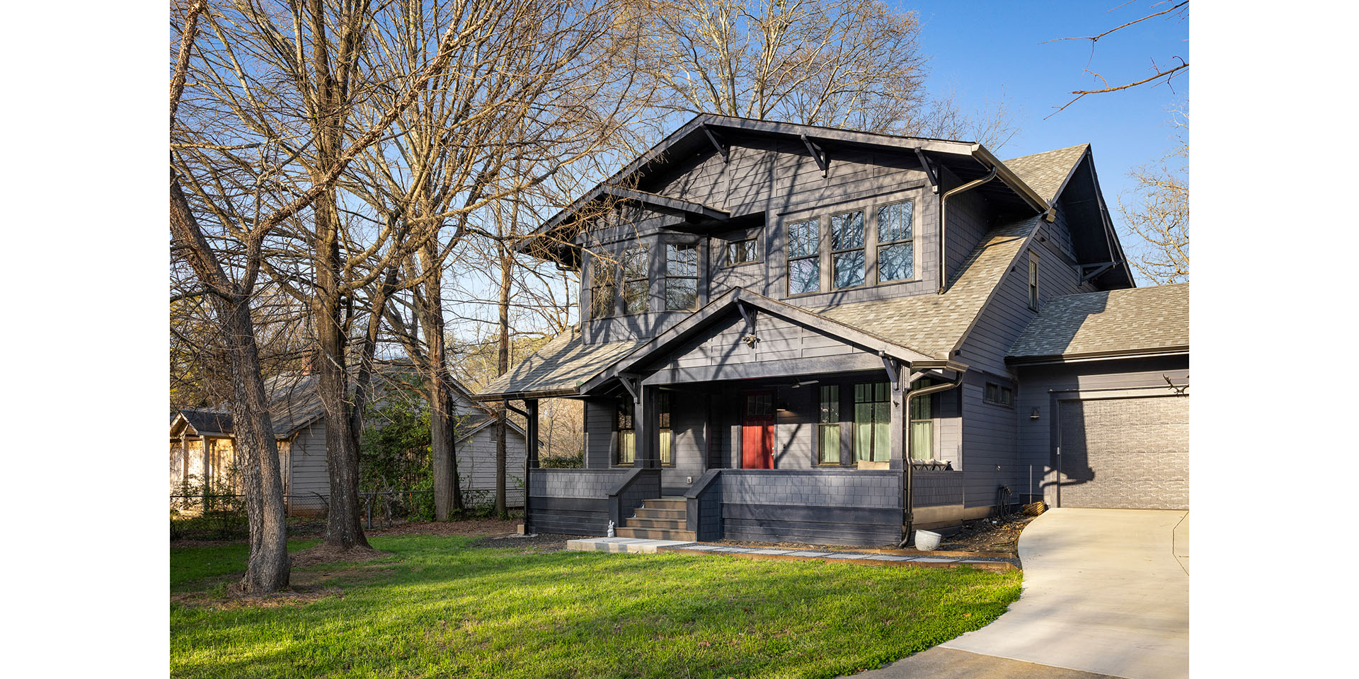 Front view of a gray, multi-gabled house with a covered porch, large windows, and a red front door, surrounded by leafless trees and green lawn.