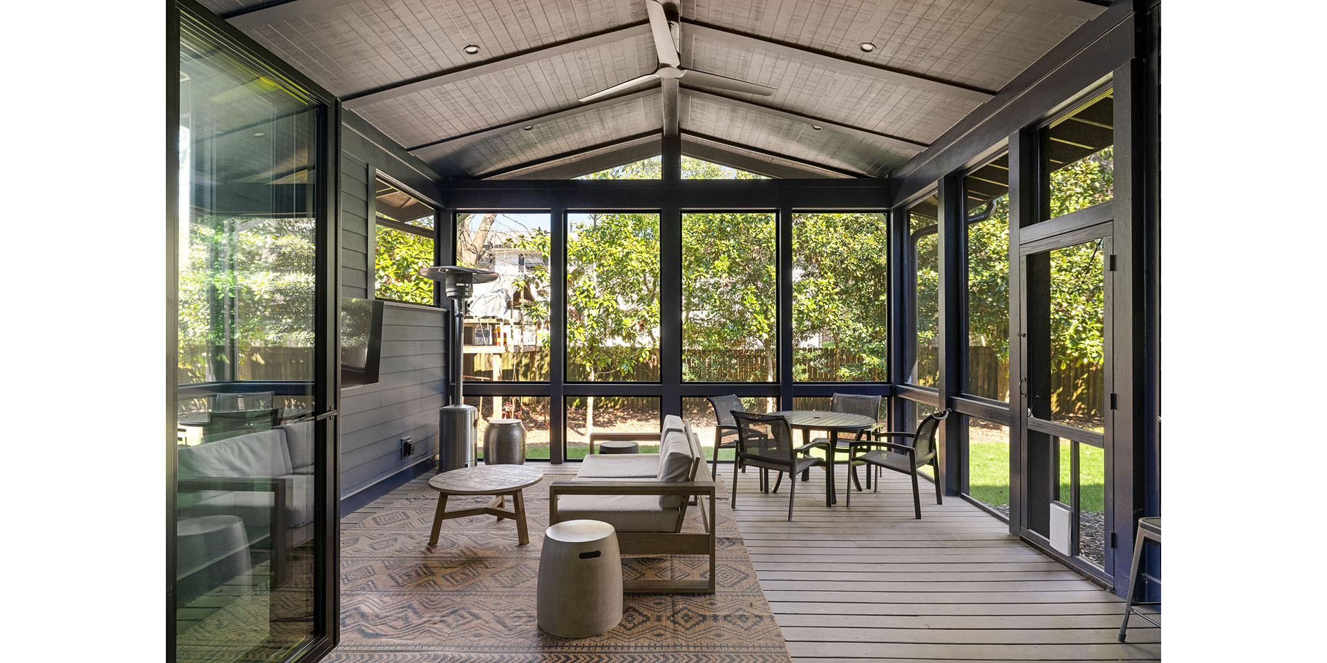 Enclosed glass-walled sunroom with dark framing, outdoor seating, and a ceiling fan overlooking a green backyard.