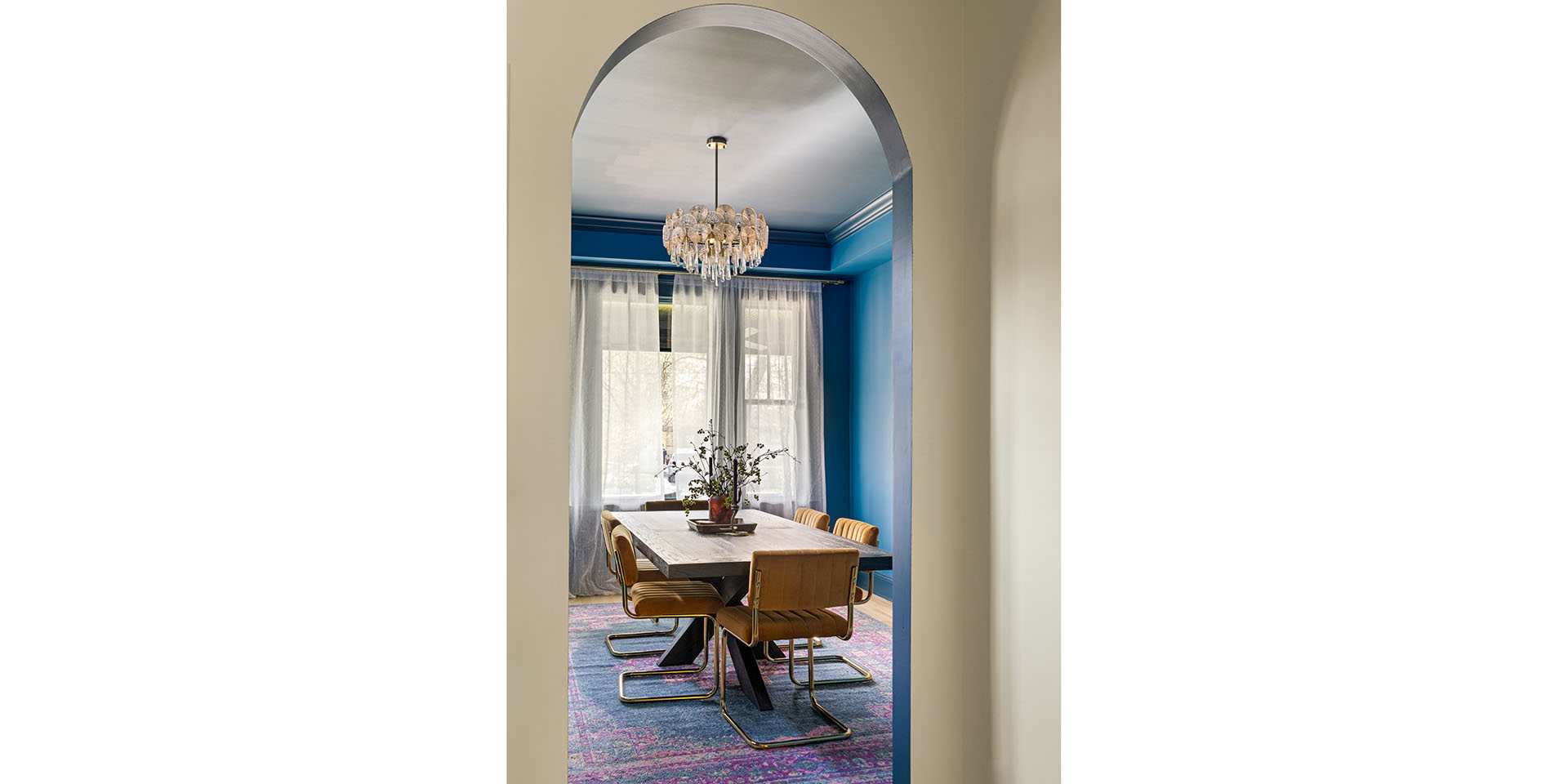 Dining room seen through an arched doorway: blue walls, crystal chandelier, sheer curtains, and a wooden table with tan chairs.