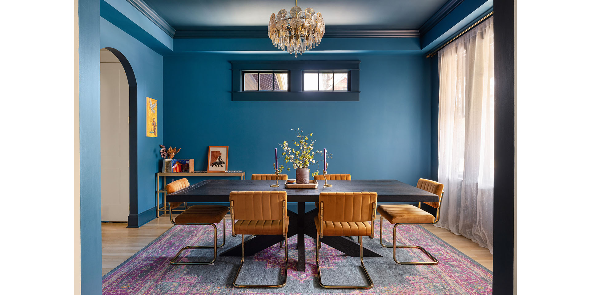 Blue dining room with a dark rectangular table and orange leather chairs under a crystal chandelier, rug, and sheer-curtained windows on the right.