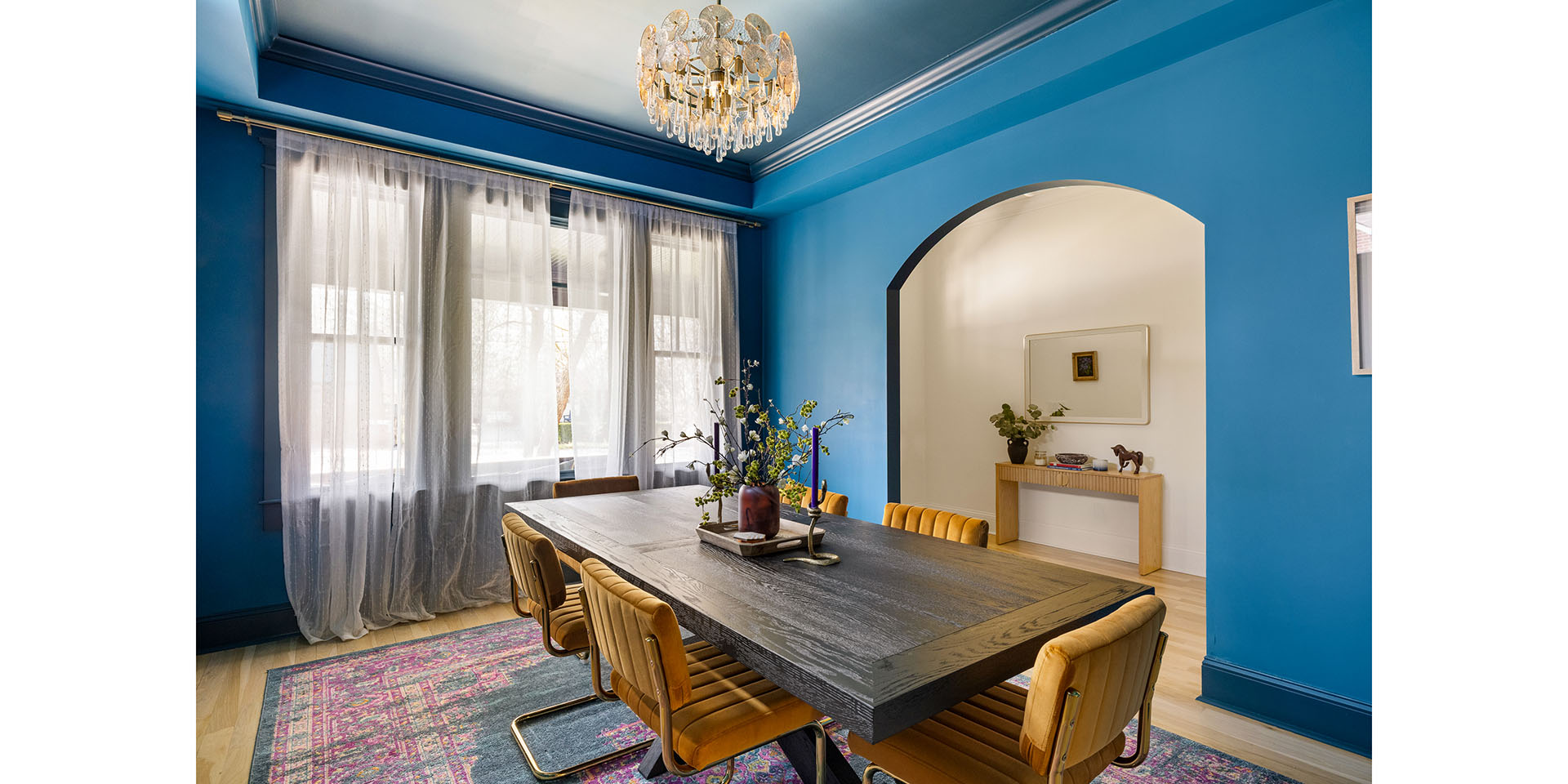 Blue dining room with a dark wooden table, tan leather chairs, and a crystal chandelier overlooking sheer-curtained windows.