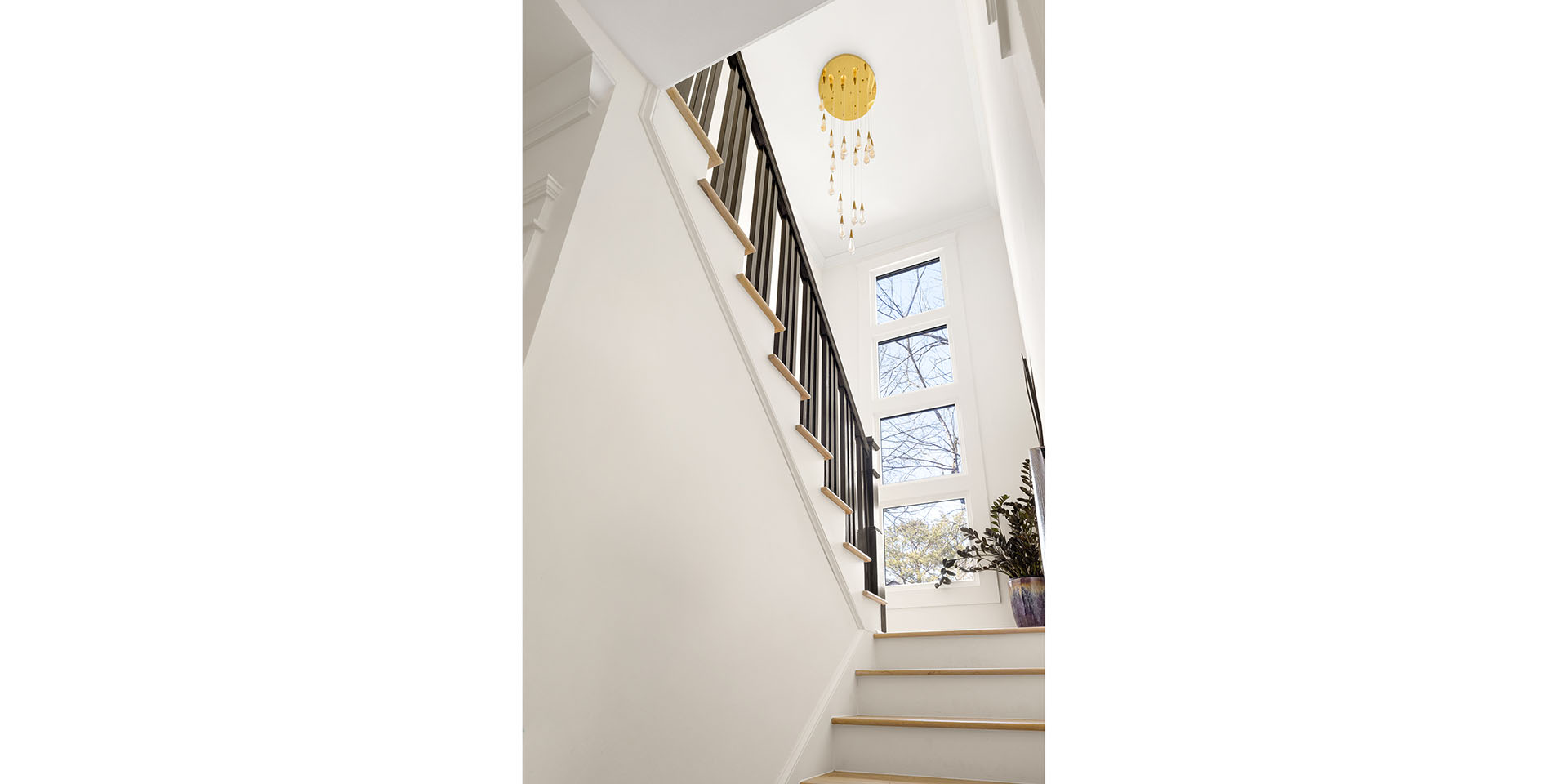 Bright white stairwell with light wood steps, black railing, large windows, and a gold chandelier hanging from the ceiling.