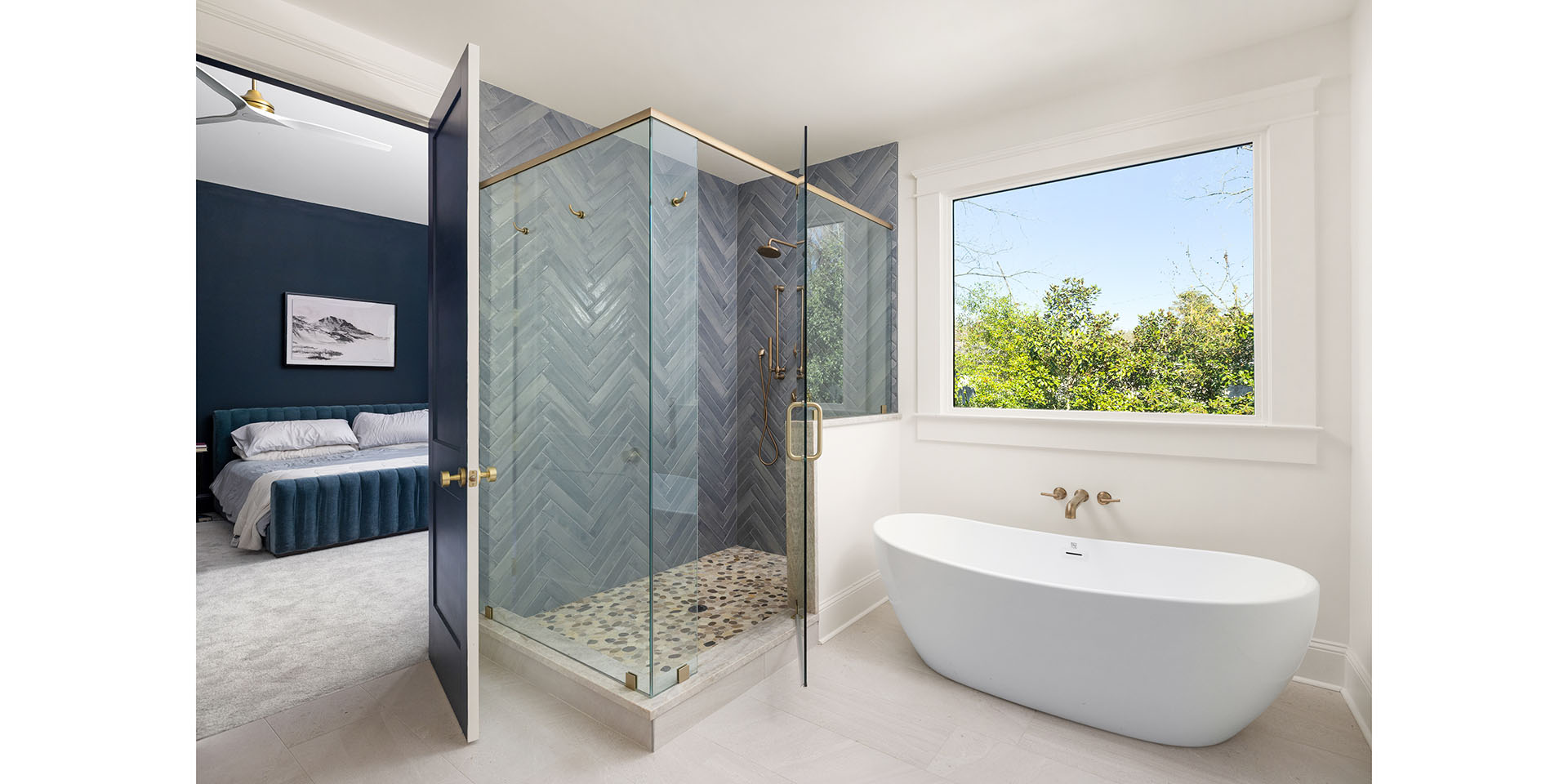 Modern ensuite bathroom with glass-walled shower in blue herringbone tile, white freestanding tub, brass fixtures, and a large window showing greenery.