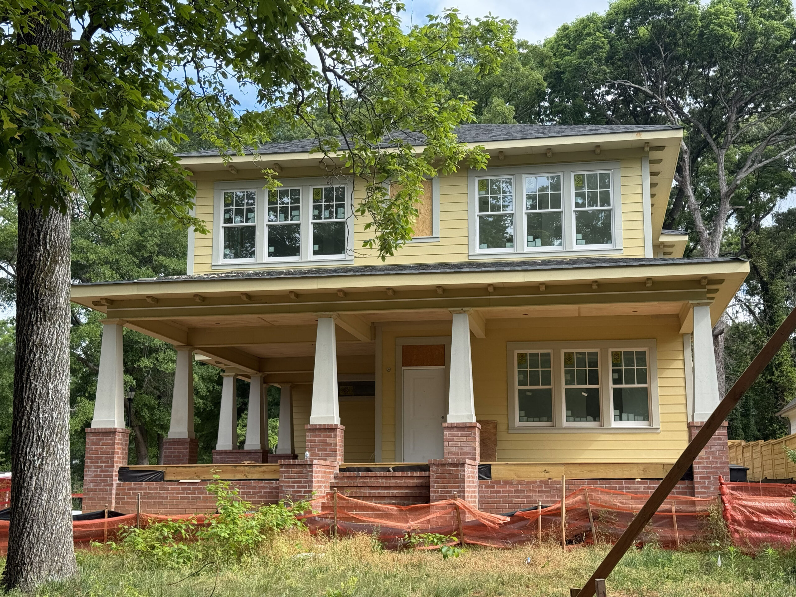 Yellow two-story house under construction with brick columns and orange safety fencing in a grassy yard, surrounded by trees