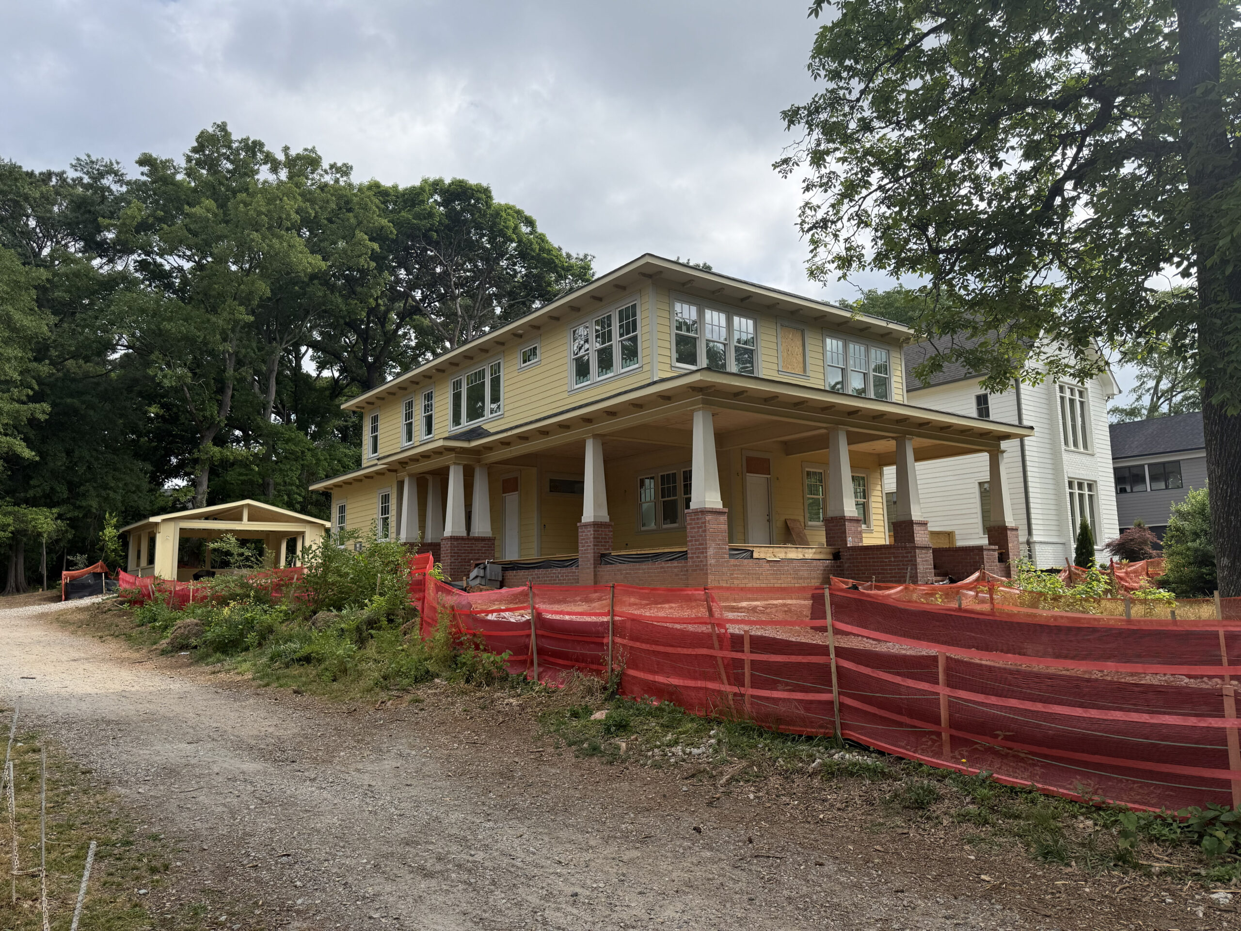 Two-story yellow house under construction with red safety fencing, brick pillars, and a gravel foreground, surrounded by mature trees.