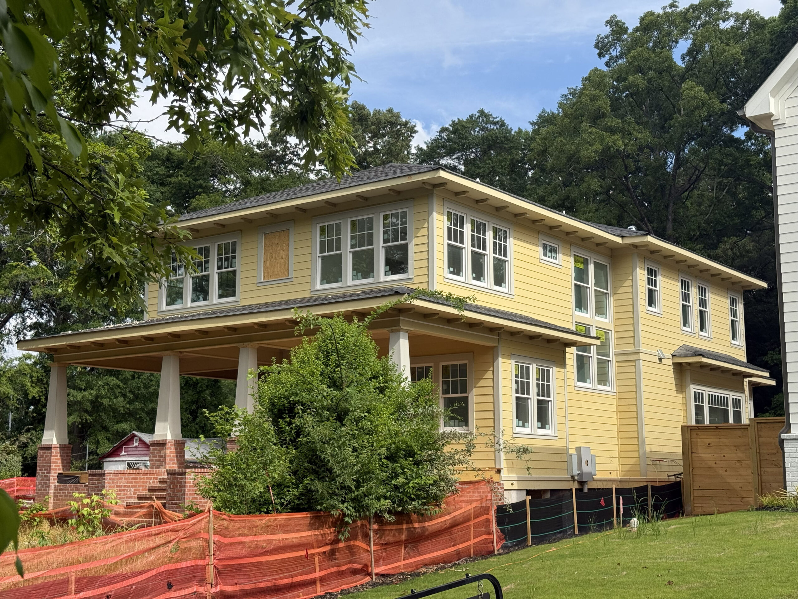 Two-story yellow house under construction with boarded window, orange safety fencing, and green trees in the background under a blue sky.